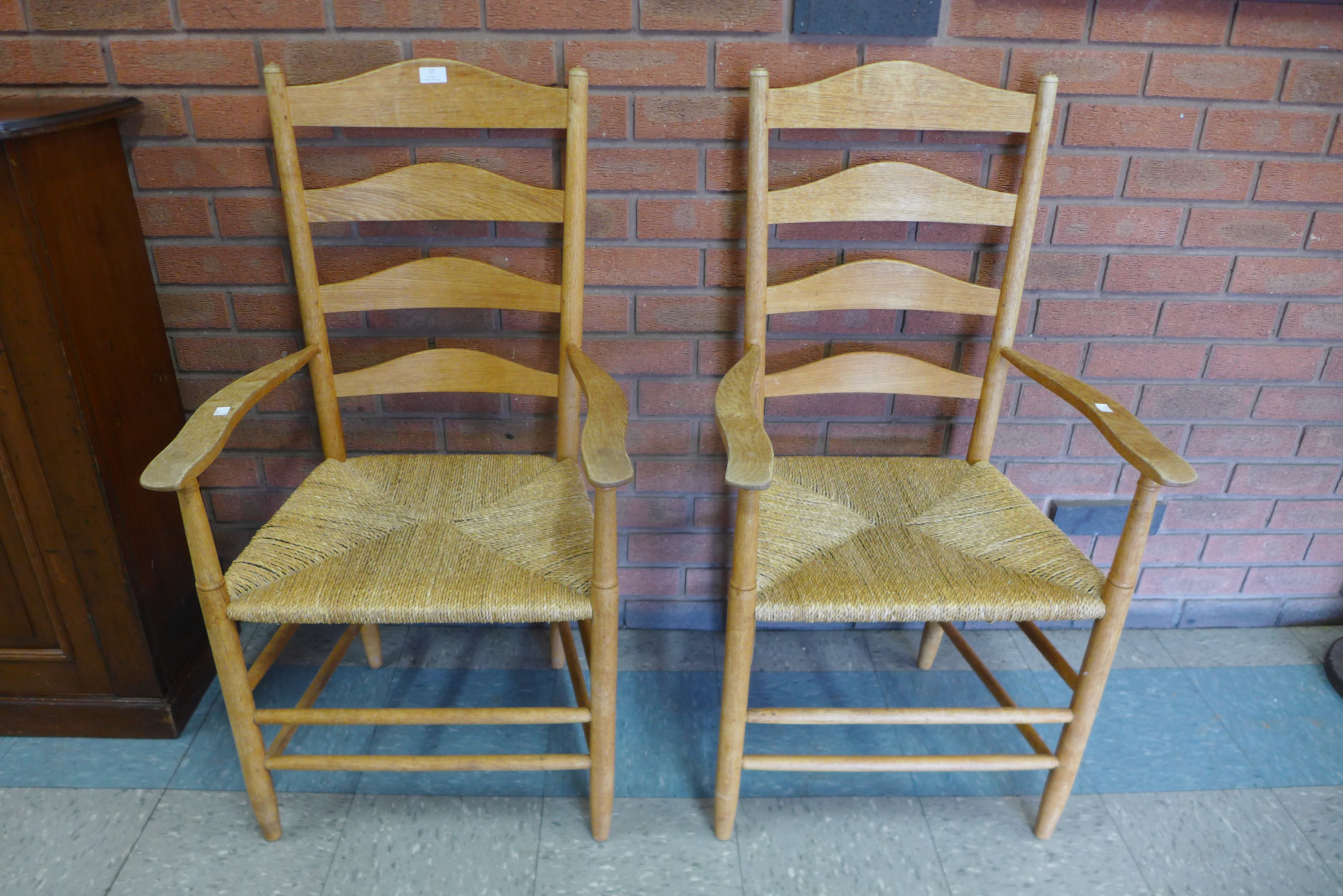 A pair of Arts and Crafts Cotswold School oak rush seated ladderback elbow chairs, attributed to