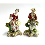 A pair of continental figurines of a gentleman and lady seated upon tree stumps, floral encrusted,
