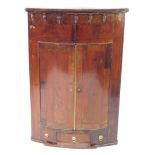 An early 19thC mahogany barrel shaped hanging corner cupboard, with carved urn top, raised above two