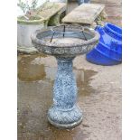 A bird bath, with a circular top on a baluster stem raised with berries and leaves on a circular rop