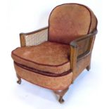 An early 20thC walnut bergere armchair, with padded back and seat, upholstered in deep red patterned