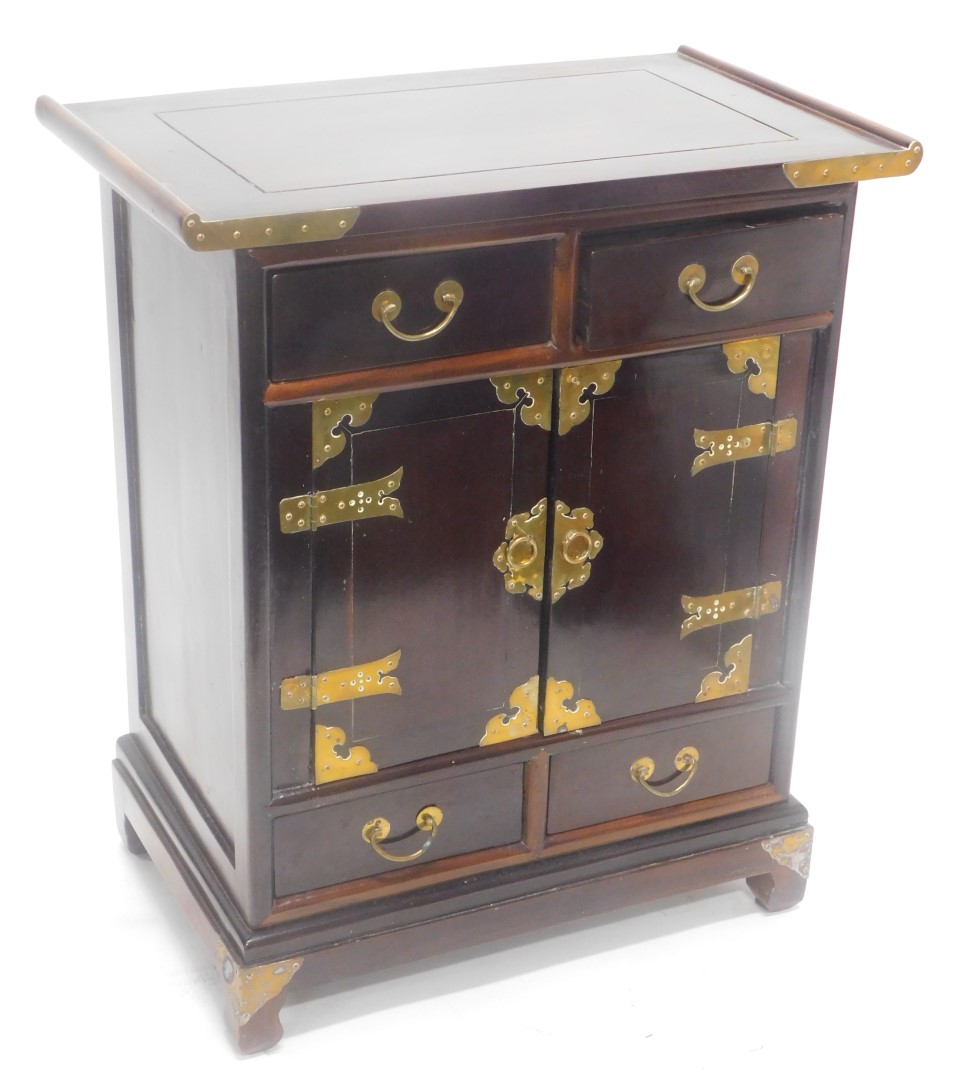 A Chinese hardwood and brass side cabinet, with a shaped top above two frieze drawers and two doors,