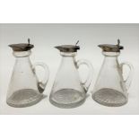 A set of three Edwardian silver hinge lidded whisky flagons with tapered glass bodies and star cut