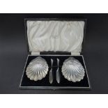 A pair of George V silver scallop shell butter dishes with a pair of butter knives, maker S.I.LD,