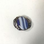 An oval blue and white polished stone within a white metal setting with leaf and vine decoration (