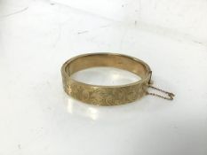 A 9ct gold bangle with clasp and safety chain, with foliate decoration to one side (6cm x 5.5cm) (