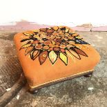 A mid 20thc footstool with a stylised flower embroidered top, on orange field, supported by bun feet