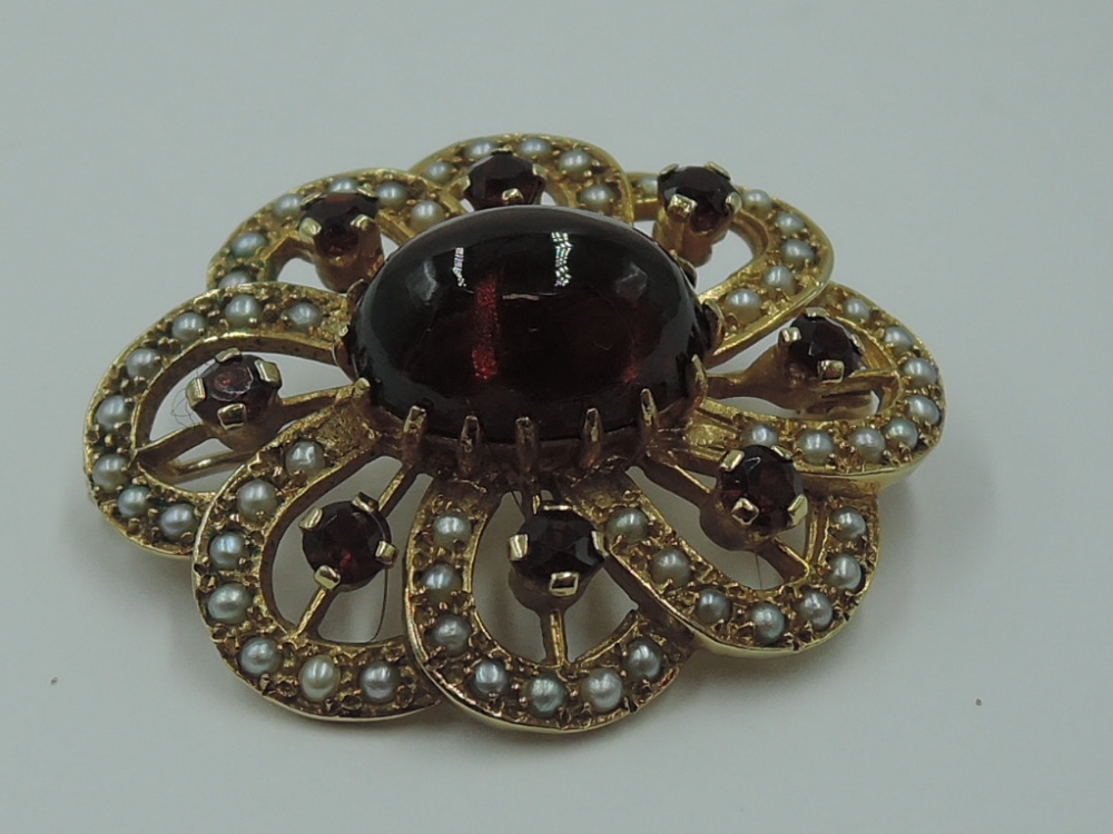 A yellow metal brooch having central garnet cabouchon within an open surround of seed pearls and