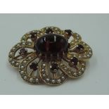 A yellow metal brooch having central garnet cabouchon within an open surround of seed pearls and