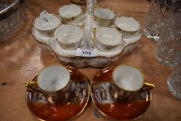 A vintage ceramic egg cup holder and two cabinet cups and saucers depicting classical scene.