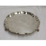 A silver salver of plain circular form having an indented pie crest rim and trefoil scroll feet,