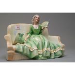 A katzhutte figurine of lady on sofa holding a fan with bonnet by her side in green and yellow