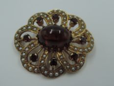A yellow metal brooch having central garnet cabouchon within an open surround of seed pearls and