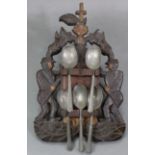 Five pewter trefid spoons (w.a.f.), mounted on a carved wood figural wall-mounted spoon rack; 15”