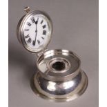 An Edwardian silver inkwell of compressed round form, the hinged lid inset timepiece to the lid