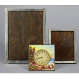 An enamelled white metal square bedside timepiece painted with a wooded lake scene, with easel