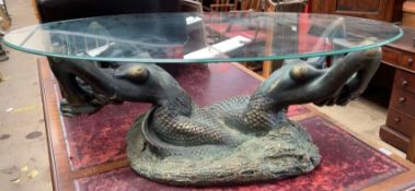 An oval glass topped coffee table held aloft by a pair of mermaids