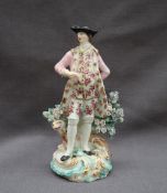 A Derby porcelain figure in a tricorn hat and pink frock coat leaning against a tree stump and