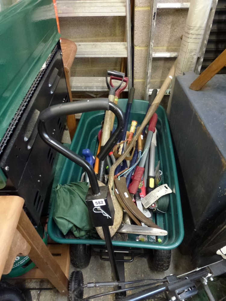 A green garden pull along trolley containing gardening tools including spades, scythes, loppers,