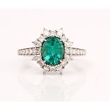 A white gold, emerald and diamond oval cluster ring, claw set with the oval cut emerald in a...