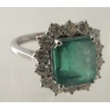 An 18ct white gold cluster ring set with an emerald cut emerald and a halo of round brilliant cut