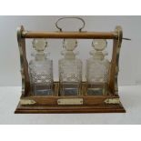 AN EARLY 20TH CENTURY OAK TANTALUS, holding three square, cut glass spirit decanters with facet