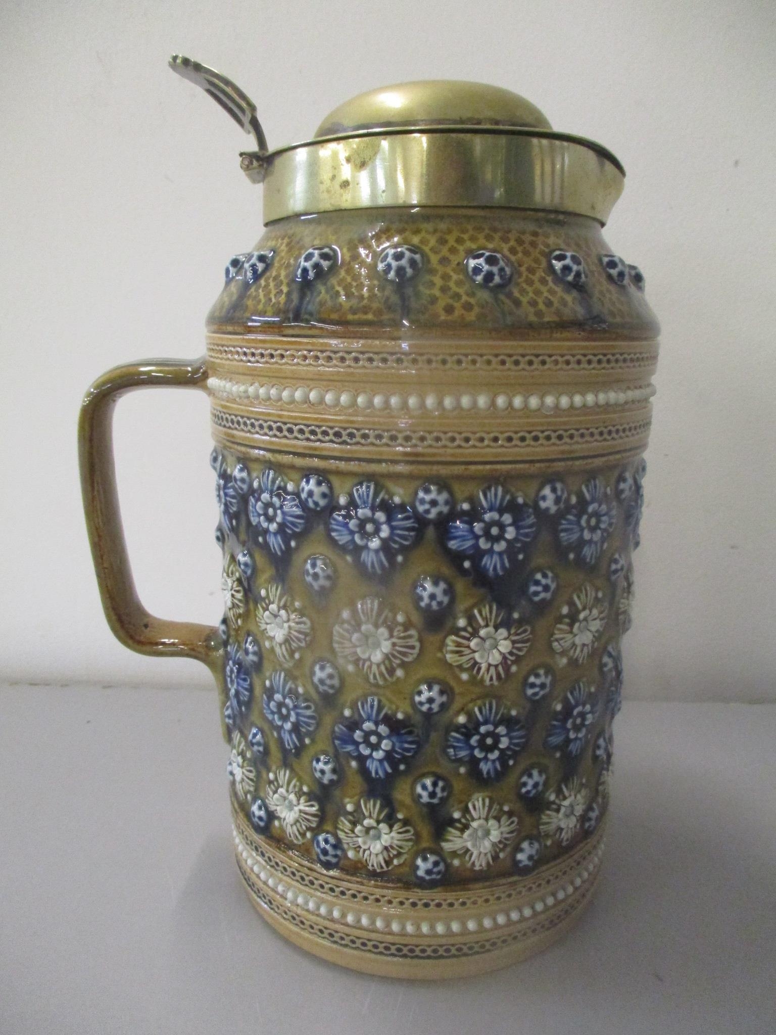 A Doulton Lambeth stoneware jug having a brass lid and decorated with raised floral detail, numbered