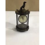 AN OMEGA MECHANICAL CLOCK WITH A DOG FINIAL TO THE TOP IN A GLASS CASE, 14CM