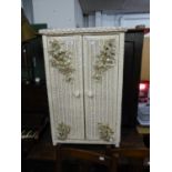 A CREAM PAINTED LLOYD LOOM STYLE CUPBOARD, THE DOORS HAVING FLORAL EMBOSSED DECORATION