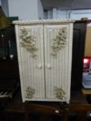 A CREAM PAINTED LLOYD LOOM STYLE CUPBOARD, THE DOORS HAVING FLORAL EMBOSSED DECORATION