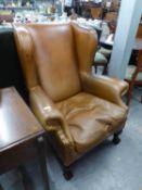 A LARGE AND HEAVY WINGED FIRESIDE ARMCHAIR COVERED IN TAN HIDE, ON CARVED MAHOGANY CABRIOLE FRONT