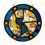 A medieval style circular stained glass panel depicting a figure by an open fire,