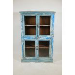 An Indian hardwood cabinet with four glazed doors and distressed paintwork, raised on a shaped