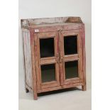 An Indian hardwood cabinet with two glazed doors, galleried top and distressed paintwork, 24" x