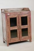 An Indian hardwood cabinet with two glazed doors, galleried top and distressed paintwork, 24" x
