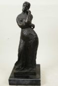 A bronze sculpture of a girl in a long dress standing in a pensive pose on a square marbled base,