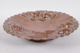 A beaten copper pedestal fruit dish with embossed apple, pear and grape decoration, 10¾" diameter