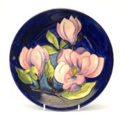 A Moorcroft plate decorated in the magnolia pattern upon a dark blue glazed ground