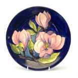 A Moorcroft plate decorated in the magnolia pattern upon a dark blue glazed ground