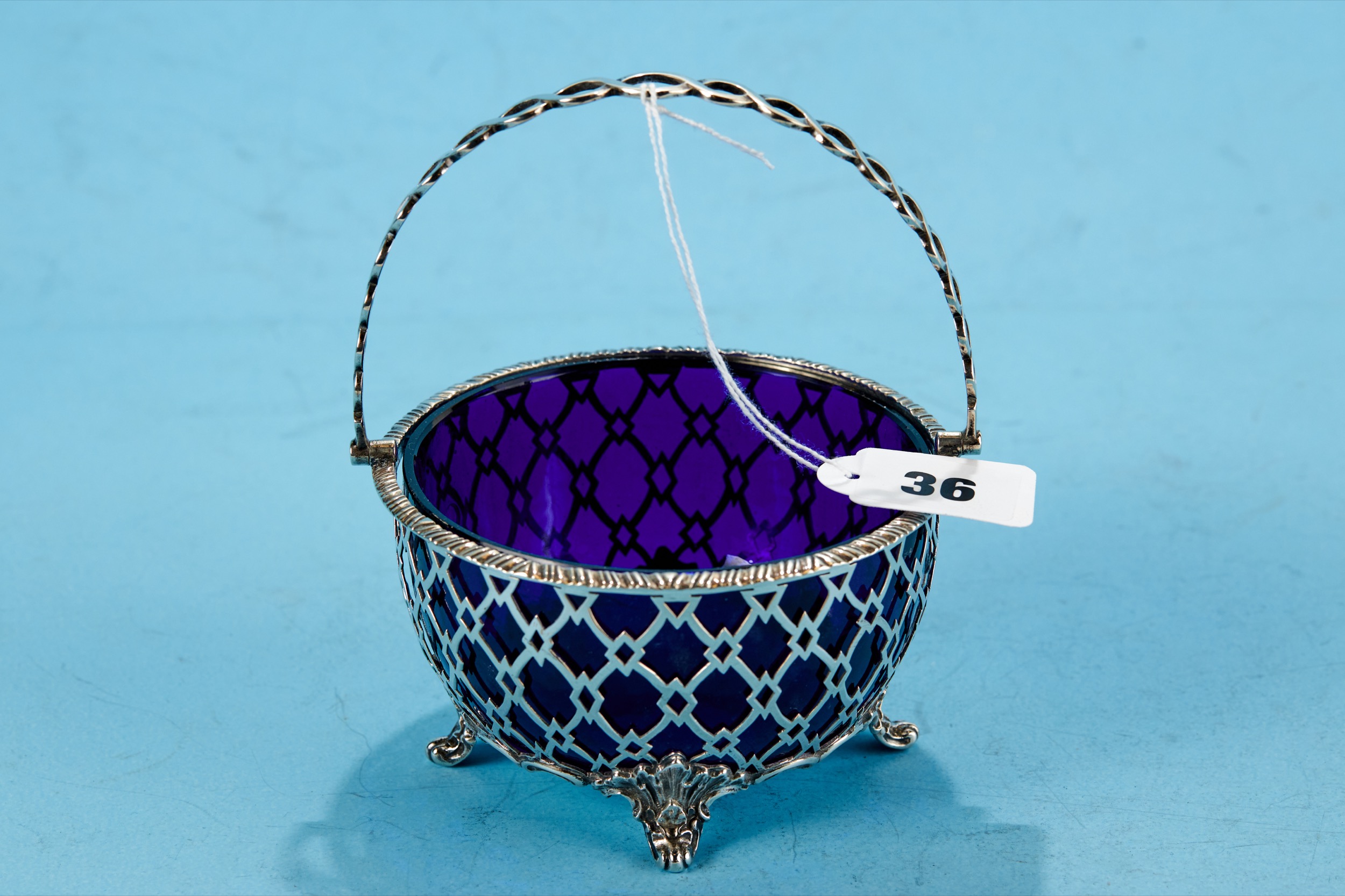 A GEORGE V CIRCULAR PIERCED SILVER SWING HANDLE SUGAR BASIN, blue glass liner, raised on scroll