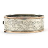 Antique unmarked silver hinged bangle engraved with faces on a wall before buildings, 6.5cm wide,
