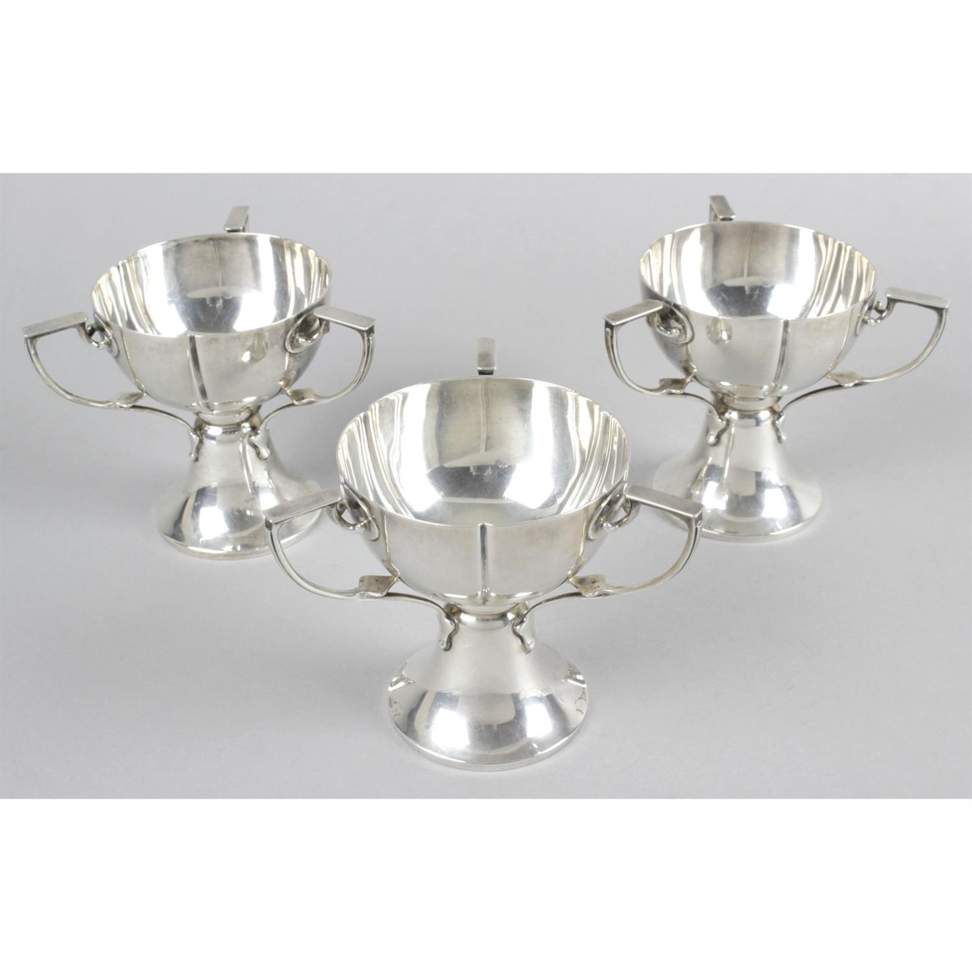 Three Edwardian silver pedestal cups, in Art Nouveau style, each with three handles. (3).
