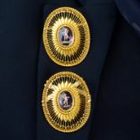 A pair of brooches, each comprising an enamel putto purple glass within a pierced radial surround.