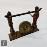 An early 20th century Malaysian gong supported by two figures in traditional dress, width 58cm.