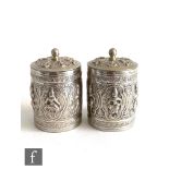 A pair of Indian white metal cylindrical spice canisters each decorated with embossed deities within