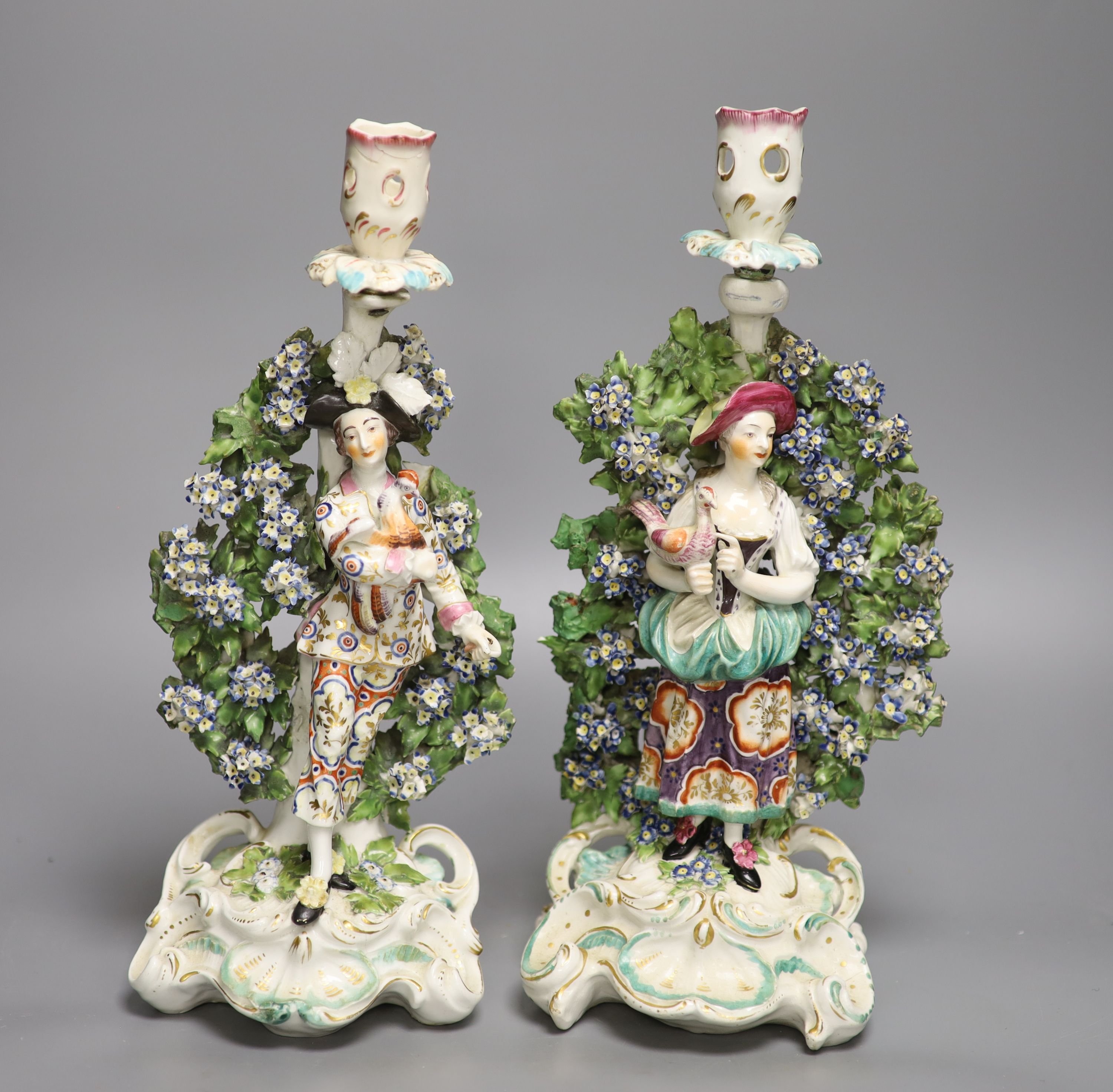 A pair of Derby candlestick figures of the Italian Farmer and wife, he holding a rooster, and her
