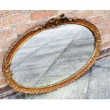 AN OVAL GILT MIRROR having a three bow carved finial, the plate measures H 48cm W 75cm, overall H