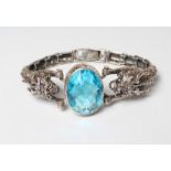 A SILVER BRACELET, the large facet cut oval turquoise glass panel open back set between two