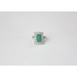 An emerald and diamond ring the step cut stone claw set in a border of fourteen round diamonds on