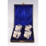 A SET OF FOUR ENGRAVED SILVER NAPKIN RINGS of shaped oval form with bun feet in a fitted case,